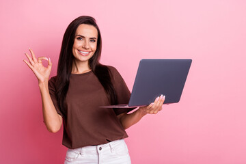 Portrait of cheerful business analytic young woman wearing brown t shirt show okey sign with netbook isolated on pink color background