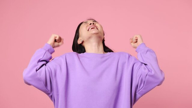 Excited jubilant overjoyed happy young woman of Asian ethnicity wear purple sweatshirt do winner gesture celebrate clench fists say yes isolated on plain pastel light pink background studio portrait