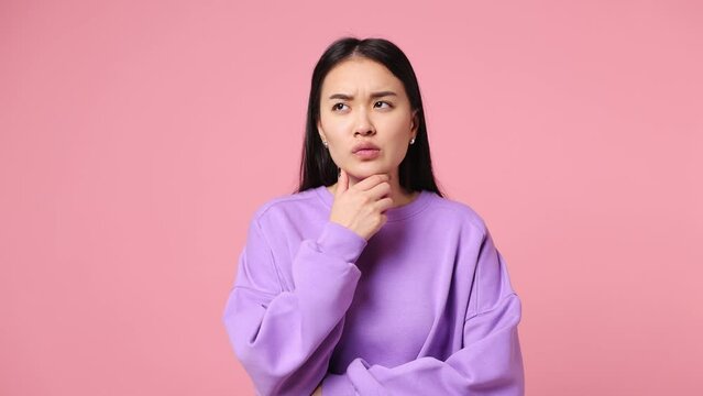 Puzzled young woman of Asian ethnicity wear sweatshirt look aside put hand prop up on chin iterates over solution options feels doubtful thinks but no decision comes isolated on plain pink background
