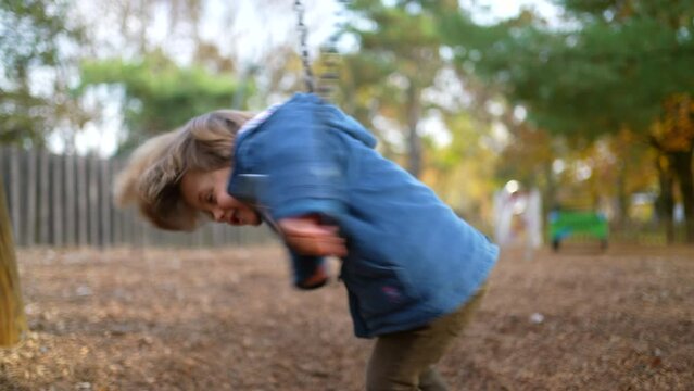 Child engaged in play outside at park swing, twisting and turning in carefree manner during solo play amidst orange autumn day foliage. Happy Little boy leaning on swing