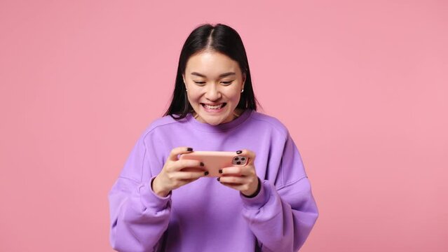 Gambling excitable young woman of Asian ethnicity wears purple sweatshirt use play racing app on mobile cell phone hold gadget smartphone for pc video games isolated on plain pastel pink background