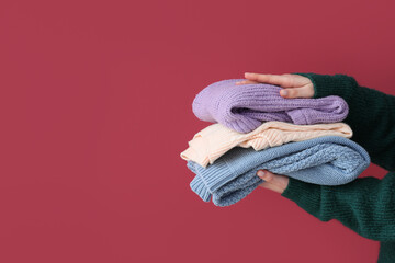 Woman holding stack of different stylish sweaters on burgundy background
