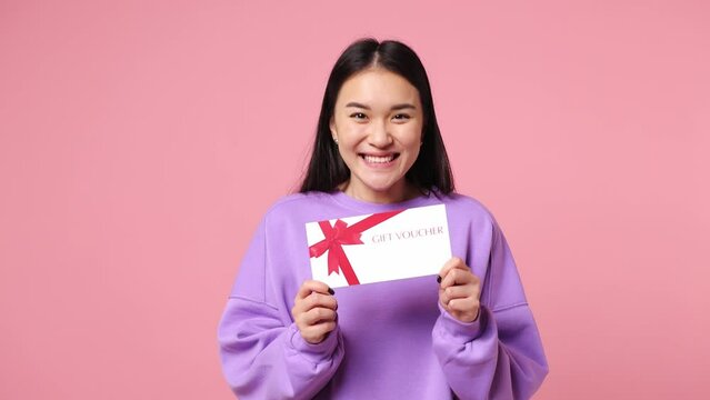 Surprised shocked happy young woman of Asian ethnicity wearing purple sweatshirt hold gift certificate coupon voucher card for store isolated on plain pastel light pink background. Lifestyle concept