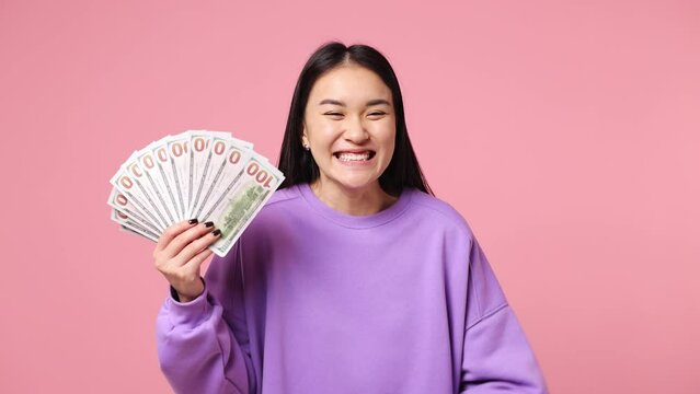 Happy young woman of Asian ethnicity wearing purple sweatshirt hold in hand fan of cash money in dollar banknotes doing winner gesture celebrate isolated on plain pastel light pink background studio