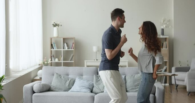 Excited attractive couple celebrating festive event, mortgage, anniversary, dancing in cozy spacious living room interior, singing song, having fun, enjoying motion, activity together
