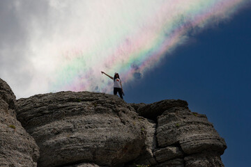A young girl at the peak of a mountain and a rainbow in the sky.