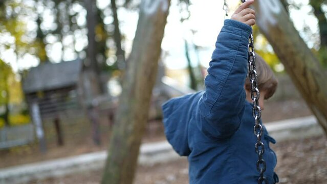 Child sitting on park swing during autumn fall day wearing blue jacket. 3 year old child seats outdoors during sunny day