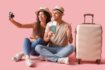 Couple of tourists with suitcase and passports taking selfie on pink background - obrazy, fototapety, plakaty
