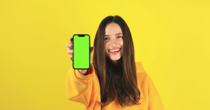 Young developer woman hold smartphone with green screen chroma key mock up recommend good application promotional sale offer, point to phone screen. Brunette girl isolated on yellow studio background.