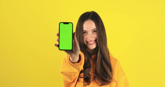 Young developer woman hold smartphone with green screen chroma key mock up recommend good application promotional sale offer, show phone screen. Brunette girl isolated on yellow studio background.