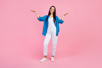 Full length photo of positive woman wear stylish shirt white pants palms demonstrate objects empty space isolated on pink color background