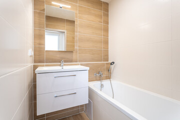 Fototapeta na wymiar modern bathroom with beige and wood-effect tiled walls, a white vanity with a sink, a large mirror, and a white bathtub with a shower attachment, illuminated by natural light from a small window