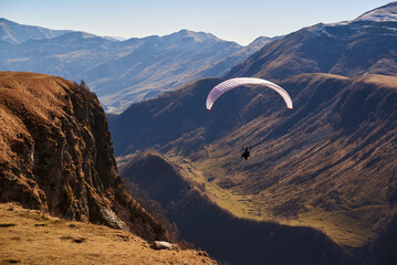 A paraglider gracefully soars above mountain peaks, their vibrant wing contrasting the blue sky,...