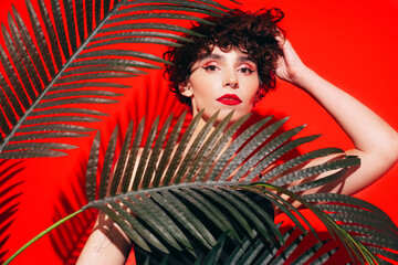 Fashion beauty portrait of young confident brunette woman with evening stylish makeup and perfect clean skin. Sexy model with curly hair posing in studio. With red bright lips. Tropical palm leaf
