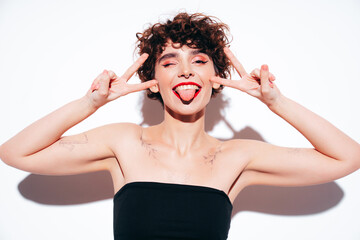Young beautiful smiling female in trendy summer black tank top. Carefree woman posing near white wall in studio with curly hairstyle. Positive model having fun, going crazy. Shows peace sign
