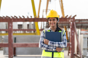 Asian woman engineer holding document smiling at construction site. Confident female Indian wearing protective helmet and vest working in factory making precast concrete wall for real estate housing.