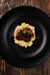 Tender beef cheeks over creamy millet, elegantly plated on a matte black dish with a rustic wooden backdrop, viewed from above