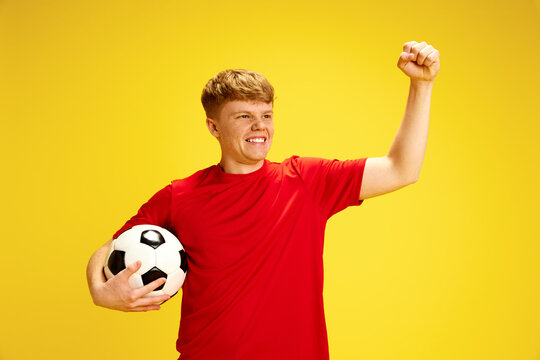Promotional material for community soccer league. Young man in red shirt holding soccer ball and making victory gesture against yellow background. Active lifestyle, youth, hobby and emotions concept