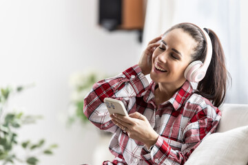 Smiling young woman sitting on couch with cell phone listening music with headphones