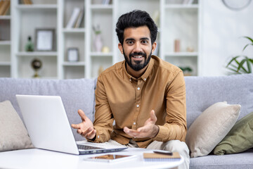Cheerful young Indian man engaging in a video chat on his laptop from the comfort of his living room sofa at home.