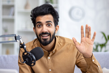 A cheerful young Indian man waving while talking into a microphone, sitting on a sofa in the cozy...