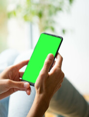 Blurred image of young woman sitting in relaxed pose. Smartphone with green screen in hands close up