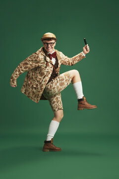 Young smiling man in image of detective, wearing retro clothes, sneaking with magnifying glass against green studio background. Concept of imagination, retro style, fashion, youth, creativity