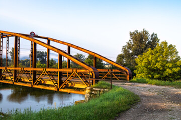 A steel bridge over a water channel connecting dirt roads. There is a sign on the bridge specifying the maximum weight limit for a passing vehicle. - obrazy, fototapety, plakaty