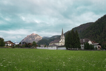 Green Meadows, Wooden Houses and Church in the Village of San Vigilio di Marebbe and Italian Alps Mountains in the Background on a Cloudy Summer Day, Italy