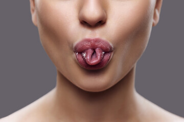 Cropped photo of. Close-up of woman's mouth with twisted tongue, highlighted by subtle lip gloss, against gray background. Concept of exercises for facial muscles, augmentation, rejuvenation treatment