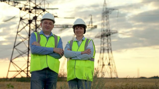Happy workers team stands crossing arms against electric power transmission lines supports in sunset field. Engineers with crossed arms rest after work at power distribution substation at sunset