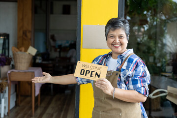 A healthy Asian senior barista in her 60s holds an open sign in front of a bakery, inviting...