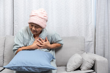 An elderly Asian woman having a heart attack sits on the sofa in the living room. The elder...