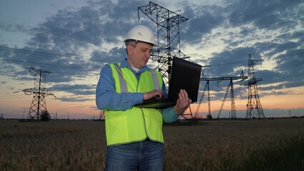 Senior engineer types on laptop standing against power transmission lines in evening rural field....