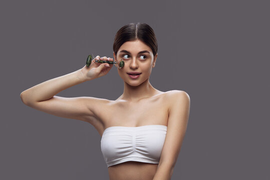 Young brunette woman using roller from natural stone, and massaging face to smooth wrinkles and prevent double chins. Concept of facial treatment and daily skincare routines using eco-friendly tools.