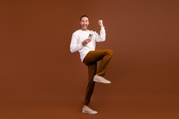 Fototapeta na wymiar Full length photo of lucky impressed guy dressed white pullover rising fist sale shopping modern device isolated brown color background