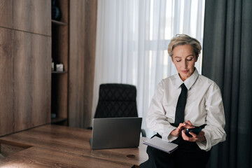 Portrait of confident middle-aged business woman using typing smartphone sitting on office desk. Female entrepreneur enjoy online chat, internet purchase, browse web, download freeware application.