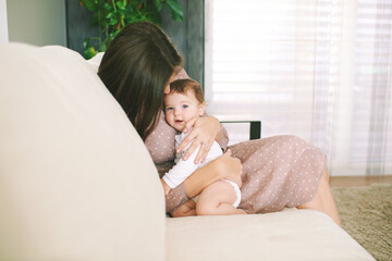 Indoor portrait of happy young mother playing with adorable baby, cuddling on white comfortable...