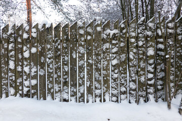 An old fence made of gray boards with pointed ends is covered with snow. White spears stuck to the surface.