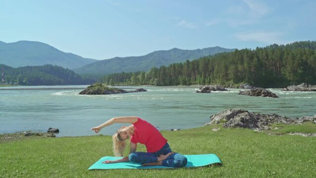 Young woman does yoga against mountain and mountain river view. Beautiful girl practices asanas and stretching muscle in morning. Exercise in nature. Meditation with view of mountains. 