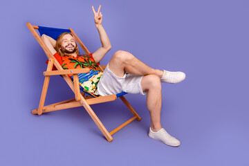 Full size photo of cheerful man wear print shirt sit on armchair showing v-sign on summer weekends isolated on purple color background
