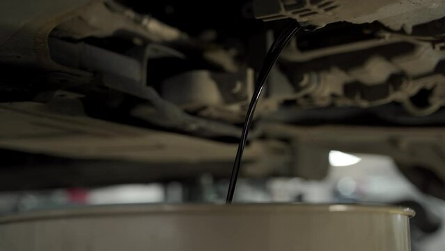 Auto mechanic drains used motor oil from car during routine service in garage. Vehicle maintenance, repair work underway. DIY car care, engine lubricant change.