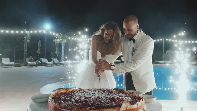 Newlyweds cut the wedding cake. Lovely bride and groom couple cutting a wedding cake dessert with a knife. Lovely man and woman make the first cut of three-tiered wedding cake. Outdoors, slow motion