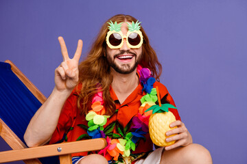 Photo of cheerful man with long hair in sunglass sit on armchair hold pineapple cocktail show v-sign isolated on violet color background