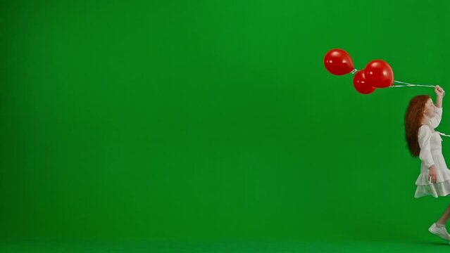 Children creative concept. Portrait of little girl in studio. Little girl in white dress with red helium balloons on green screen. The girl is jumping into the frame.