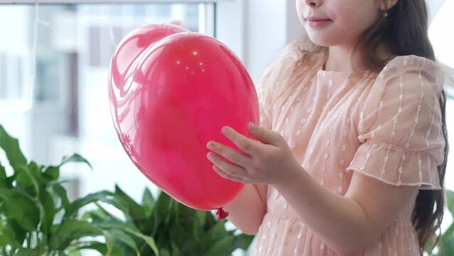 A 9-year-old little girl in a peach dress holds and twirls a large red balloon in the shape of a heart in her hands close-up. valentine's day