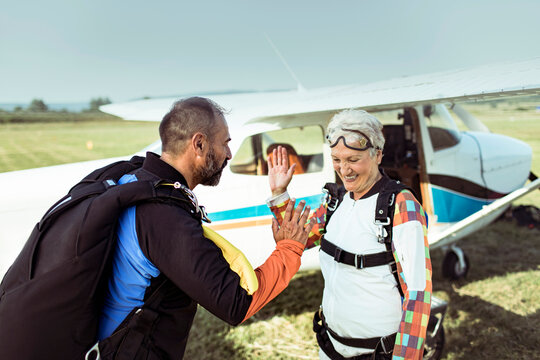 Happy senior woman and skydiving instructor celebrating successful jump with airplane in background