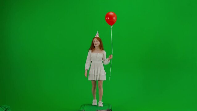 Little girl in white dress with red balloon on chroma key green screen isolated background in party hat walking looking at the camera smiling.