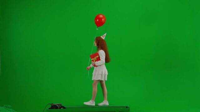 Redheaded little girl in white dress with a festive cap on her head and with a gift in her hands is walking with a red balloon. Green screen. Side view. Concept of holiday, joy and fun.