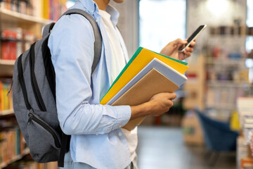 Black male student holds copybooks and using cellphone in library, showcasing harmonious blend of...